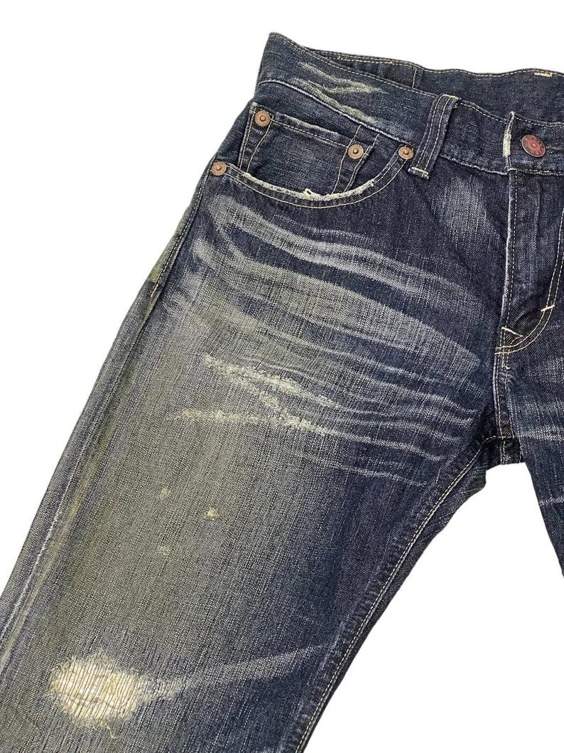Puede incluir: Primer plano de vaqueros de mezclilla azul desgastados. Los vaqueros tienen un lavado descolorido con desgaste visible, incluyendo parches claros y un &aacute;rea desgastada cerca de la rodilla. Incluye remaches met&aacute;licos y un dise&ntilde;o cl&aacute;sico de cinco bolsillos.