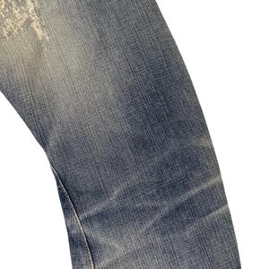Puede incluir: Primer plano de una secci&oacute;n de vaqueros azules. Los vaqueros muestran signos de desgaste con zonas descoloridas y un parche de color claro cerca del bolsillo. La tela tiene un aspecto texturizado.