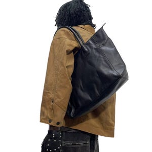 May include: Black leather tote bag with double handles, carried over the shoulder. The bag is rectangular and appears to be empty. The person is wearing a brown jacket and black jeans.