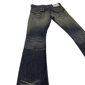 Puede incluir: Vaqueros de mezclilla de lavado oscuro con dise&ntilde;o de pierna acampanada. Los vaqueros presentan el estilo cl&aacute;sico de cinco bolsillos, con costuras decorativas en los bolsillos traseros. Una peque&ntilde;a etiqueta es visible en la cintura trasera. Los vaqueros est&aacute;n planos sobre un fondo blanco.