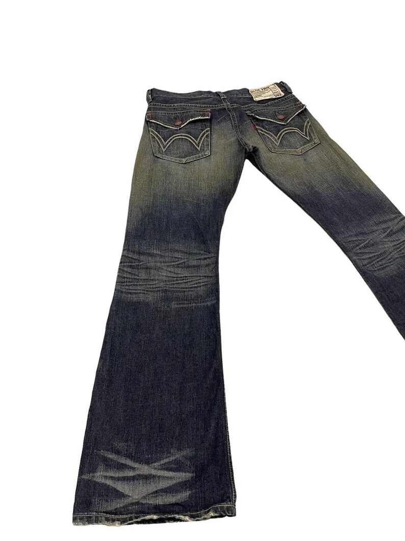 Puede incluir: Vaqueros de mezclilla de lavado oscuro con dise&ntilde;o de pierna acampanada. Los vaqueros presentan el estilo cl&aacute;sico de cinco bolsillos, con costuras decorativas en los bolsillos traseros. Una peque&ntilde;a etiqueta es visible en la cintura trasera. Los vaqueros est&aacute;n planos sobre un fondo blanco.