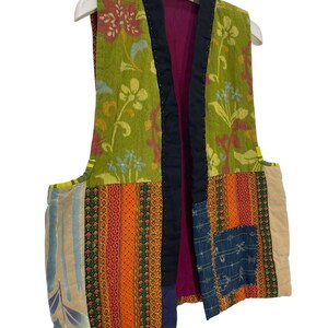 May include: A patchwork vest with a floral pattern in shades of green, pink, and yellow. The vest features a black trim and a magenta lining. The lower section has a mix of patterned fabrics in orange, blue, and beige.