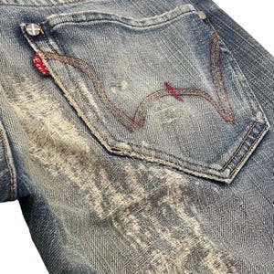 Puede incluir: Primer plano de unos vaqueros de mezclilla azules con un lavado desgastado. El bolsillo trasero presenta costuras rojas en un dise&ntilde;o distintivo. Una peque&ntilde;a etiqueta roja con texto blanco es visible, junto con un parche de cuero rojo en la parte superior.