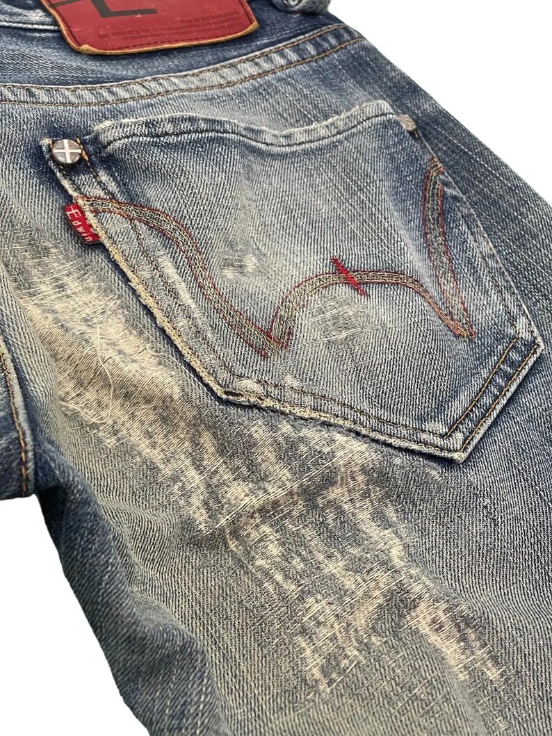 Puede incluir: Primer plano de unos vaqueros de mezclilla azules con un lavado desgastado. El bolsillo trasero presenta costuras rojas en un dise&ntilde;o distintivo. Una peque&ntilde;a etiqueta roja con texto blanco es visible, junto con un parche de cuero rojo en la parte superior.
