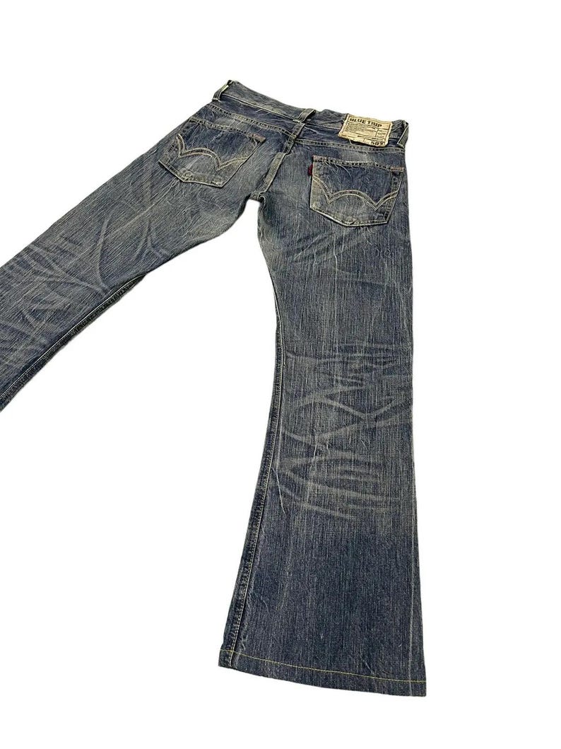 May include: Faded blue denim jeans with a flared leg design. The jeans have a worn appearance with visible fading and distressing. Features include classic five-pocket styling, a red tag, and a leather patch on the back. The jeans are laid flat against a white background.