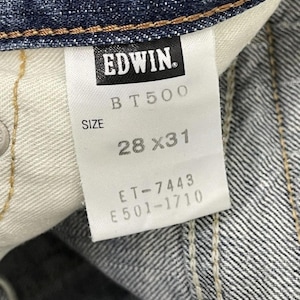 May include: Close-up of a denim jeans tag with the brand name "EDWIN" in black. The tag also displays "BT500", "SIZE 28X31", and other product codes. The jeans are a dark blue wash.