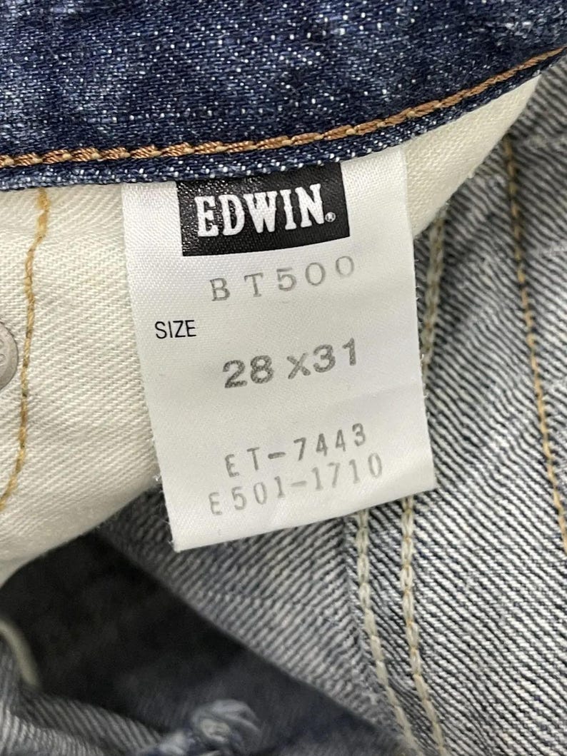 May include: Close-up of a denim jeans tag with the brand name "EDWIN" in black. The tag also displays "BT500", "SIZE 28X31", and other product codes. The jeans are a dark blue wash.