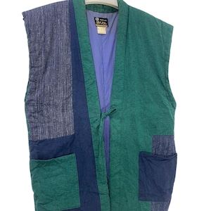 May include: A sleeveless, quilted vest in shades of green, blue, and purple. The vest features a patchwork design with two front pockets and a tie closure. The interior lining is a light purple color. The vest is hanging on a white hanger.