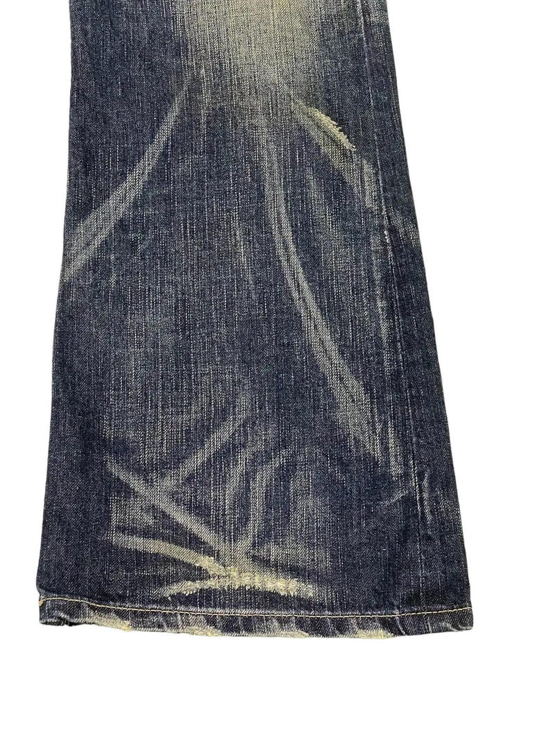 Puede incluir: Primer plano de una secci&oacute;n de vaqueros de mezclilla azul oscuro con detalles blancos descoloridos y desgastados. Los vaqueros tienen un aspecto desgastado con vetas de color claro y un dobladillo deshilachado, lo que sugiere un estilo vintage o pre-usado.