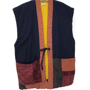 May include: A sleeveless navy blue jacket with a patchwork design. The jacket features a mustard yellow lining, rust-colored trim, and a tie closure. The lower section has red and gray patterned patches.