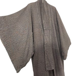 May include: A long, brown, woven robe with a textured pattern. The robe has a wide collar and long sleeves. It is hanging on a hanger.