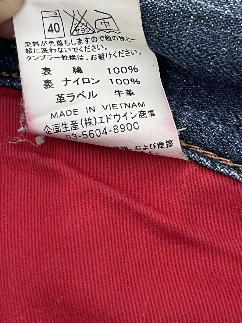 Puede incluir: Primer plano de una etiqueta de ropa con texto en japon&eacute;s e ingl&eacute;s. La etiqueta est&aacute; adherida a tela vaquera y tela roja. La etiqueta incluye instrucciones de lavado, composici&oacute;n de la tela y el texto "MADE IN VIETNAM".