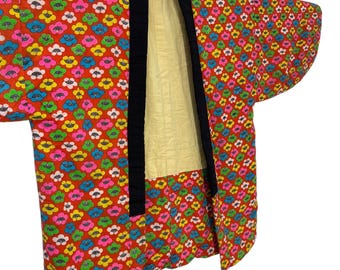 May include: A vintage kimono with a vibrant floral pattern in red, pink, yellow, blue, and green. The kimono features black trim and a light yellow quilted lining. The garment is suspended by a black strap, showcasing its design and construction.