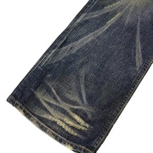Puede incluir: Primer plano de una secci&oacute;n de vaqueros azules con detalles desgastados. Los vaqueros tienen zonas descoloridas y parches m&aacute;s claros y desgastados, creando un aspecto vintage. Las costuras son visibles a lo largo de los bordes.