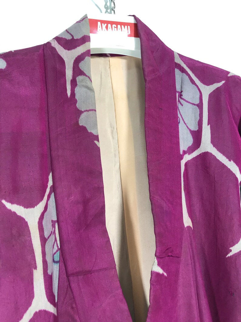 May include: A close-up of the collar of a pink kimono with a white floral pattern. The kimono is hanging on a hanger with the label "AKAGAMI" visible.