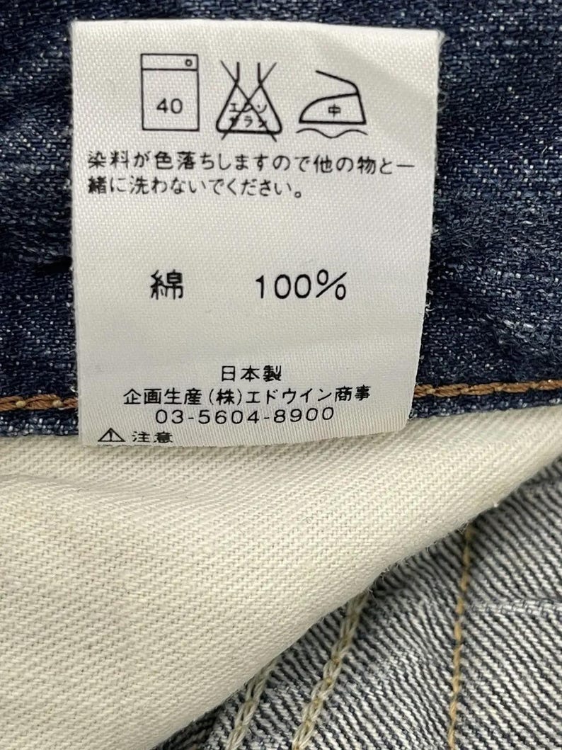 May include: A white clothing tag with care symbols and Japanese text. The tag indicates a 40-degree wash, and the fabric is 100% cotton. The tag also includes the manufacturer's information.