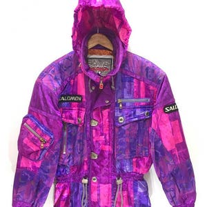 May include: A purple and pink ski jacket with a geometric pattern. The jacket has a hood, a zip closure, and multiple pockets. The brand name "Salomon" is visible on the jacket.