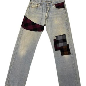 May include: Light blue denim jeans with plaid patches in burgundy, black, and gray. The jeans have a button fly and a classic five-pocket design. The patches are sewn onto the front of the jeans.