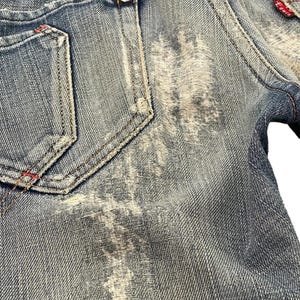 Puede incluir: Primer plano de unos vaqueros de mezclilla azul desgastados. Los vaqueros presentan un efecto de lavado descolorido, costuras visibles y una peque&ntilde;a etiqueta roja. Los bolsillos traseros tienen costuras decorativas y remaches met&aacute;licos.