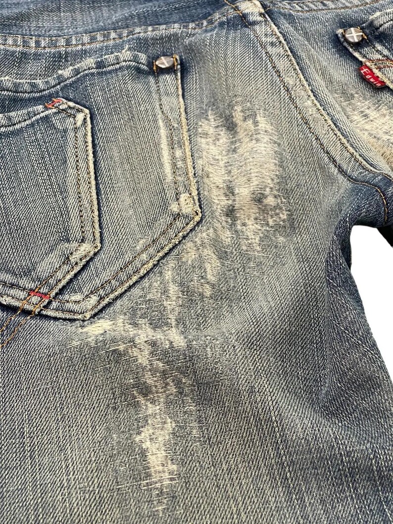 Puede incluir: Primer plano de unos vaqueros de mezclilla azul desgastados. Los vaqueros presentan un efecto de lavado descolorido, costuras visibles y una peque&ntilde;a etiqueta roja. Los bolsillos traseros tienen costuras decorativas y remaches met&aacute;licos.