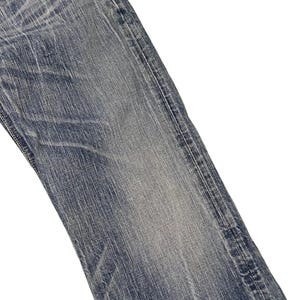 May include: Close-up of a section of blue denim jeans. The fabric shows wear and fading, with visible texture and subtle variations in color. The jeans have a classic five-pocket design.