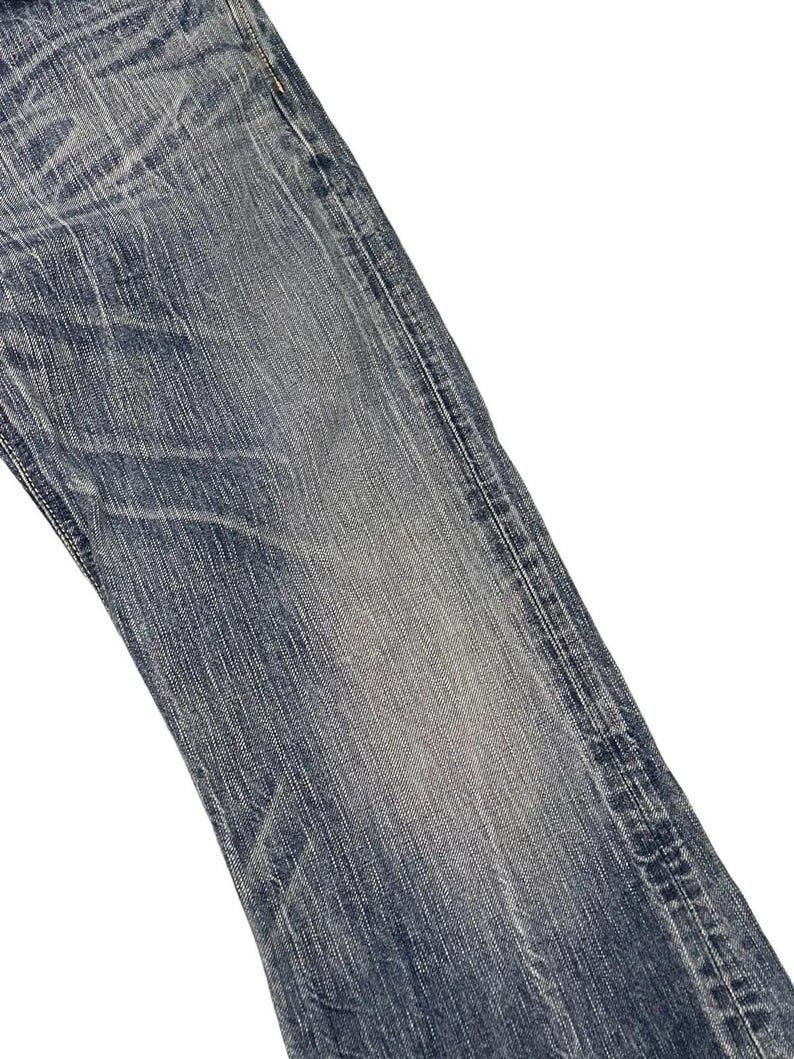 May include: Close-up of a section of blue denim jeans. The fabric shows wear and fading, with visible texture and subtle variations in color. The jeans have a classic five-pocket design.