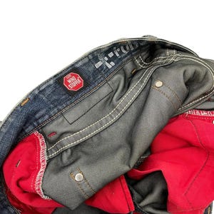 Puede incluir: Primer plano de unos vaqueros de mezclilla azules con forro interior rojo. Los vaqueros tienen costuras blancas y un parche rojo "WIND STOPPER". El interior es visible, mostrando la construcci&oacute;n y los materiales.