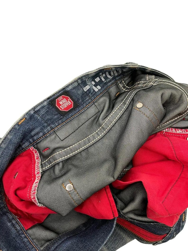 Puede incluir: Primer plano de unos vaqueros de mezclilla azules con forro interior rojo. Los vaqueros tienen costuras blancas y un parche rojo "WIND STOPPER". El interior es visible, mostrando la construcci&oacute;n y los materiales.