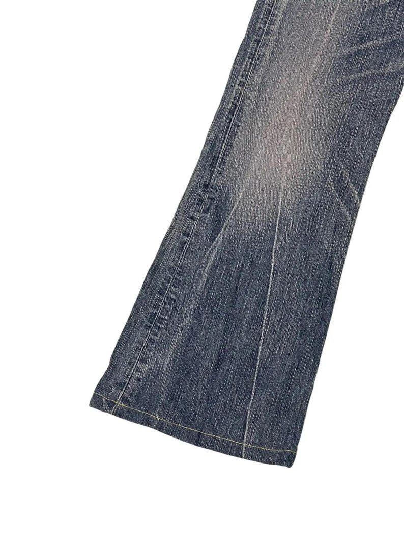 May include: Close-up of a section of blue denim jeans. The jeans have a faded wash effect, with lighter areas creating a worn appearance. Visible are the seams and stitching, with a classic five-pocket design. The denim is a medium weight.