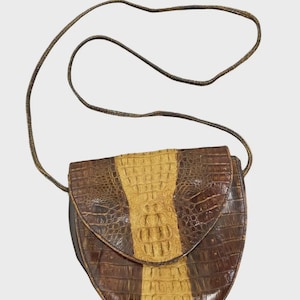 May include: A crossbody bag featuring a textured, embossed pattern resembling reptile skin. The bag has a rounded shape with a flap closure and a long, thin shoulder strap. The design incorporates a vertical light tan center panel flanked by dark brown side panels.