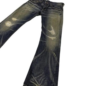 Puede incluir: Vaqueros de mezclilla de lavado oscuro con una apariencia desgastada y descolorida. Los vaqueros tienen una abertura de pierna acampanada y muestran desgaste con marcas de color claro. Los vaqueros est&aacute;n en &aacute;ngulo sobre un fondo blanco.