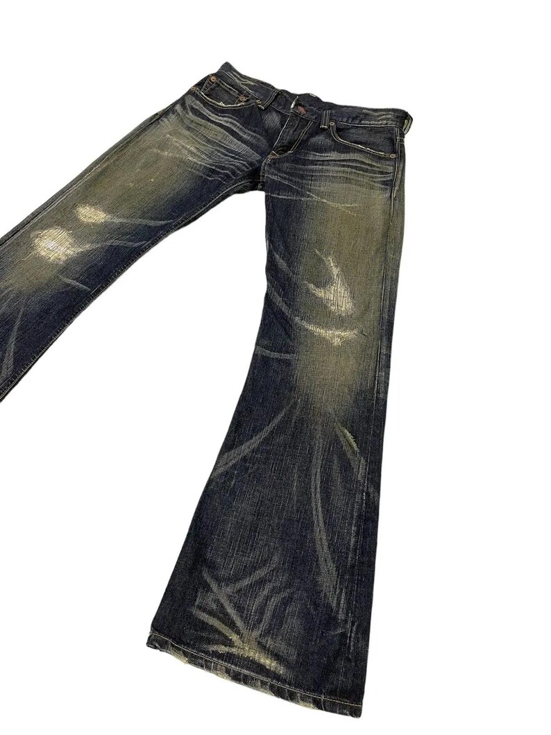 Puede incluir: Vaqueros de mezclilla de lavado oscuro con una apariencia desgastada y descolorida. Los vaqueros tienen una abertura de pierna acampanada y muestran desgaste con marcas de color claro. Los vaqueros est&aacute;n en &aacute;ngulo sobre un fondo blanco.