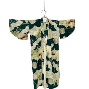 May include: A vintage kimono featuring a dark green backdrop adorned with white cranes and floral motifs. The garment, suspended from a metal hanger, showcases long sleeves and a cream-coloured tie. The kimono's length is approximately 120 cm.