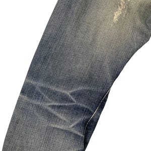 Puede incluir: Primer plano de unos vaqueros de mezclilla azules con zonas descoloridas y desgastadas. Los vaqueros muestran signos de desgaste, con parches y arrugas m&aacute;s claros que a&ntilde;aden a su est&eacute;tica vintage. La costura es visible en algunos lugares.
