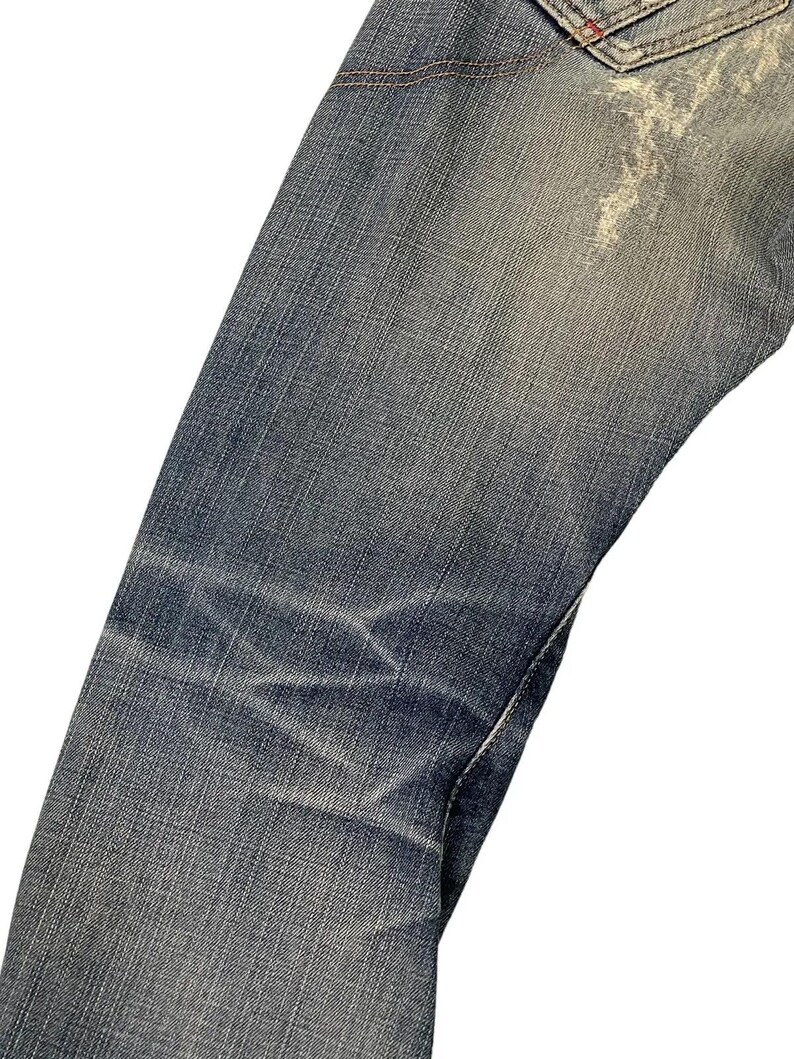 Puede incluir: Primer plano de unos vaqueros de mezclilla azules con zonas descoloridas y desgastadas. Los vaqueros muestran signos de desgaste, con parches y arrugas m&aacute;s claros que a&ntilde;aden a su est&eacute;tica vintage. La costura es visible en algunos lugares.