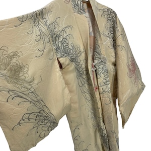 Può includere: Un kimono color crema caratterizzato da un motivo ripetuto di grandi fiori di crisantemo stilizzati in tonalità grigie e rosa tenue. Il capo presenta maniche ampie e fluide e una chiusura frontale con un piccolo cordino annodato.