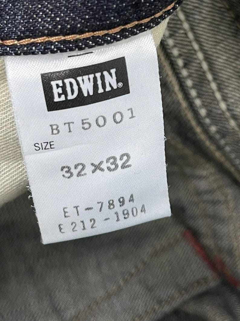 Puede incluir: Primer plano de una etiqueta blanca de vaqueros Edwin. La etiqueta muestra el nombre de la marca "EDWIN" en un rect&aacute;ngulo negro. Debajo, las palabras "BT 5001" y "SIZE". La etiqueta indica una talla de 32x32 pulgadas (81x81 cm).