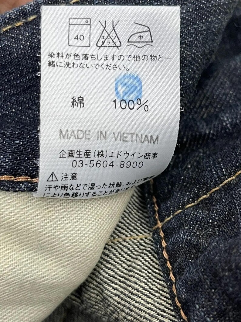 Puede incluir: Primer plano de una etiqueta blanca con texto japon&eacute;s y s&iacute;mbolos de cuidado, adherida a una tela vaquera azul oscuro. La etiqueta indica que la tela es 100% algod&oacute;n y est&aacute; hecha en Vietnam. Tambi&eacute;n se incluyen instrucciones de lavado.