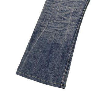 May include: Close-up of a section of blue denim jeans. The jeans have a classic five-pocket design with a straight leg. The denim has a faded, worn look with visible creases and subtle variations in color.