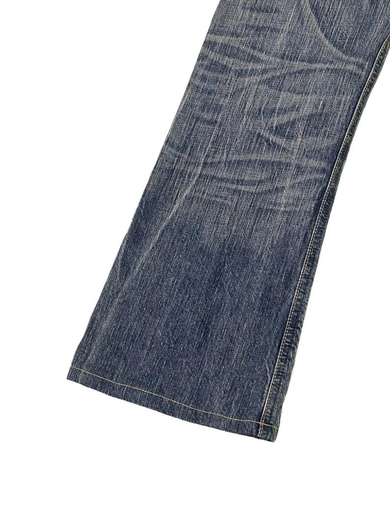 May include: Close-up of a section of blue denim jeans. The jeans have a classic five-pocket design with a straight leg. The denim has a faded, worn look with visible creases and subtle variations in color.