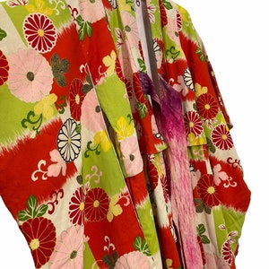 May include: A red, green, and white kimono with a floral pattern. The kimono is hanging on a hanger and has a pink sash tied around it.