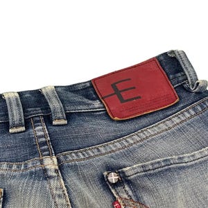 Puede incluir: Primer plano de vaqueros de mezclilla azul con un lavado descolorido. Se ve un parche de cuero rojo con un logotipo "E" negro. El bolsillo trasero tiene un bot&oacute;n plateado y una etiqueta roja Edwin.