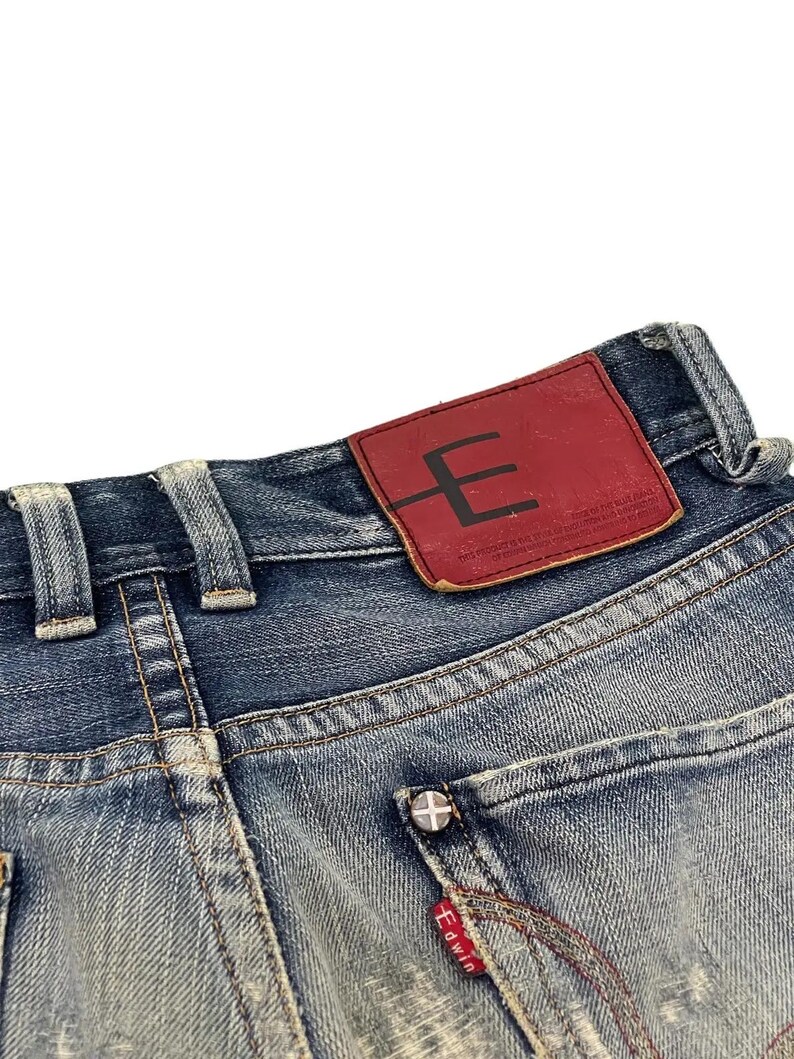 Puede incluir: Primer plano de vaqueros de mezclilla azul con un lavado descolorido. Se ve un parche de cuero rojo con un logotipo "E" negro. El bolsillo trasero tiene un bot&oacute;n plateado y una etiqueta roja Edwin.