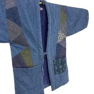 May include: A blue quilted jacket with patchwork details. The jacket features various fabric patterns in shades of blue, green, and purple. It has two front pockets and a tie closure. The jacket is hanging on a wooden hanger.