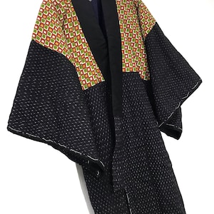 May include: A black and green patterned kimono with a black collar and a white stitching detail. The kimono is hanging on a hanger.