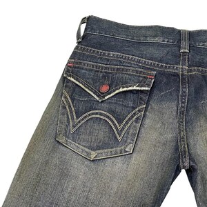 Puede incluir: Primer plano del bolsillo trasero de unos vaqueros azules. El bolsillo tiene un dise&ntilde;o &uacute;nico con costuras blancas y un bot&oacute;n rojo. Los vaqueros muestran signos de desgaste, con un aspecto descolorido.