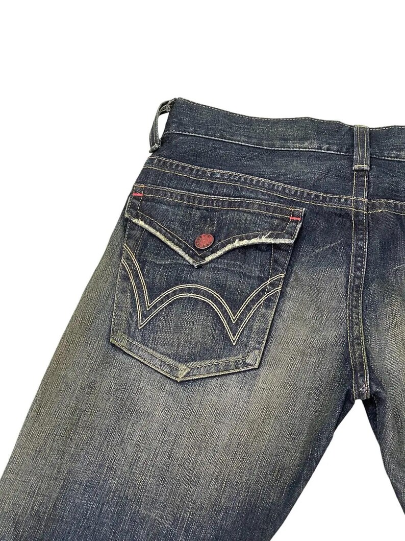Puede incluir: Primer plano del bolsillo trasero de unos vaqueros azules. El bolsillo tiene un dise&ntilde;o &uacute;nico con costuras blancas y un bot&oacute;n rojo. Los vaqueros muestran signos de desgaste, con un aspecto descolorido.