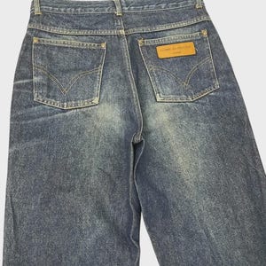 May include: Back view of blue denim jeans with two back pockets and a brown leather label. The jeans have a faded wash and a classic five-pocket design. The label reads "COMME des GARÇONS HOMME".