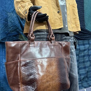 Könnte beinhalten: Eine braune Ledertasche mit einer vorderen Tasche mit Schlangenlederstruktur. Die Tasche hat zwei Griffe und zwei Seitentaschen. Die Tasche wird von einer Person mit schwarzen Handschuhen gehalten.