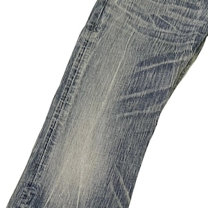 May include: Close-up of a pair of blue denim jeans. The jeans have a faded wash with visible wear and tear details. The fabric shows vertical and horizontal lines, creating a textured appearance. The jeans are angled, showing the front of the leg.
